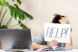 An overwhelmed woman sits at a table in front of a laptop, her head in one hand; in the other she holds a sign that reads “HELP!” The sign fully obscures her face.